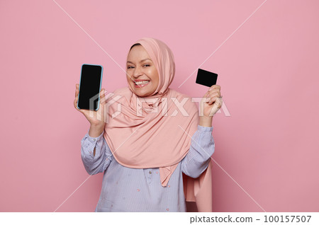 Charming Muslim woman in elegant pink hijab, smiling looking at camera, holding a smartphone and black credit card, isolated over pink color background. People Business Finance Mobile internet banking Charming Muslim woman in elegant pink hijab, smiling looking at camera, holding a smartphone and black credit card, isolated over pink color background. People Business Finance Mobile internet banking 100157507