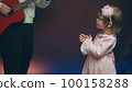 Little girl on stage in pink vintage dress, she claps his hands and dances, her father plays acoustic guitar. Color music is shining. Performance on stage. Early development of children 100158288