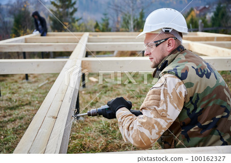 Man building wooden frame house on pile foundation. Male worker drills hole by electric drill in timber framing of future house. Builder wearing white helmet and protective goggles. Carpentry concept. 100162327