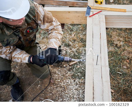 Man building frame house on pile foundation. Close up of male worker drilling hole by electric drill in wooden frameg of future house. Builder wearing white helmet. Carpentry concept. 100162329