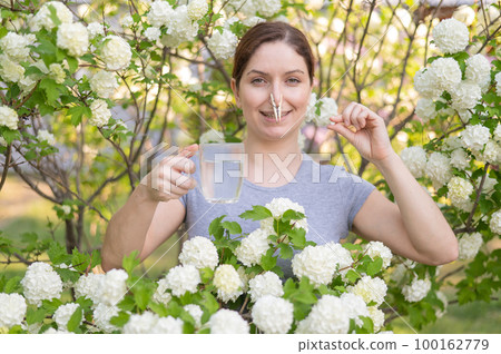 Caucasian woman takes an antihistamine medicine and removes a clothespin from her nose near a flowering tree. Caucasian woman takes an antihistamine medicine and removes a clothespin from her nose near a flowering tree. 100162779