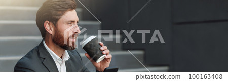 Attractive office worker in suit sitting on stairs with coffee during break and using phone 100163408