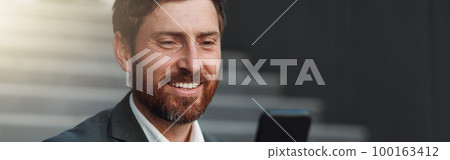 Close up of smiling office worker in suit sitting on stairs with coffee during break and using phone 100163412