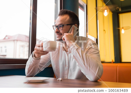 Stylish positive European guy businessman in glasses and white shirt, with a beard, talking on a smartphone and drinking coffee sitting at a table in a cafe Stylish positive European guy businessman in glasses and white shirt, with a beard, talking on a smartphone and drinking coffee sitting at a table in a cafe 100163489