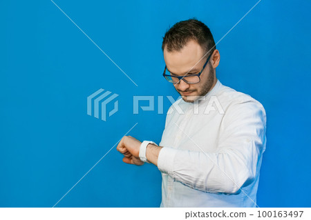 Stylish, business-like young portrait guy Caucasian-looking businessman in glasses and white shirt, looks time on smart clock on blue background 100163497