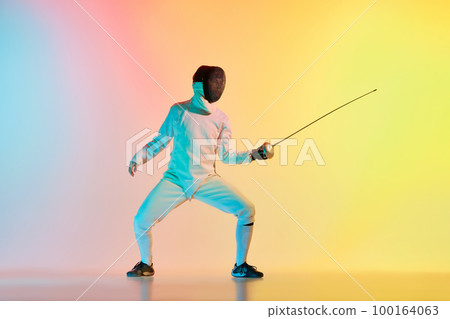 Young man, male fencer with sword practicing in fencing over gradient pink-yellow background in neon light. Sportsman shows fencing technique 100164063