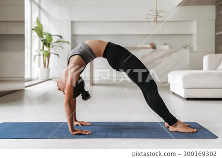 Young sporty woman practicing yoga at home on mat in Bridge pose Young sporty woman practicing yoga at home on mat in Bridge pose 100169302