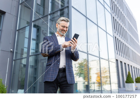 Happy senior gray-haired businessman, lawyer, investor stands outside the office, skyscraper. Joyful uses the phone, reads, sends, received. Happy senior gray-haired businessman, lawyer, investor stands outside the office, skyscraper. Joyful uses the phone, reads, sends, received. 100177257