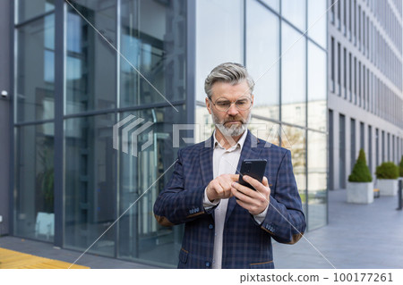 An older gray-haired man in a suit and glasses stands outside an office center, holding and using a mobile phone. He is waiting for an appointment, calls a taxi, makes phone calls. An older gray-haired man in a suit and glasses stands outside an office center, holding and using a mobile phone. He is waiting for an appointment, calls a taxi, makes phone calls. 100177261