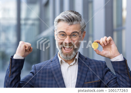 Close-up photo. A senior man, businessman standing outside office and holding bitcoin, gold coin, cryptocurrency in hand. He looks at the camera. shows a winning gesture with his hands, smiles. Close-up photo. A senior man, businessman standing outside office and holding bitcoin, gold coin, cryptocurrency in hand. He looks at the camera. shows a winning gesture with his hands, smiles. 100177263