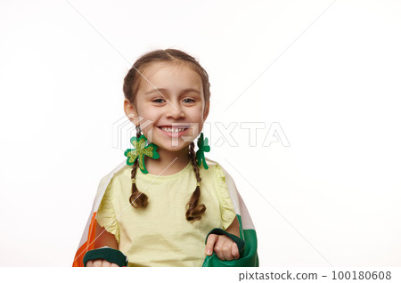 Adorable beautiful cheery Irelandish child girl with two pigtails and clover leaves earrings, carrying ireland flag and smiling at camera, isolated on white background. Saint Patrick's Day celebration Adorable beautiful cheery Irelandish child girl with two pigtails and clover leaves earrings, carrying ireland flag and smiling at camera, isolated on white background. Saint Patrick's Day celebration 100180608