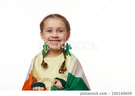 Close-up portrait of a cherry baby girl with clover leaves earring, smiling a toothy smile looking at camera, posing wrapped in flag of Ireland for Saint Patrick's Day, on isolated white background Close-up portrait of a cherry baby girl with clover leaves earring, smiling a toothy smile looking at camera, posing wrapped in flag of Ireland for Saint Patrick's Day, on isolated white background 100180609