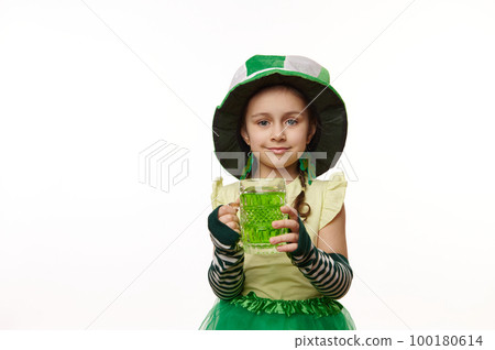 Lovely child, little girl dressed as Leprechaun in a hat with clover leaves, smiling and holding out at camera a green drink in mug, while celebrating Saint Patrick's Day, isolated on white background 100180614