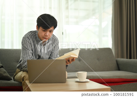 Relaxed asian man holding notebook and watching webinar or working online on laptop at home 100181591