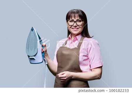 Woman in apron posing with iron, on gray background Woman in apron posing with iron, on gray background 100182233