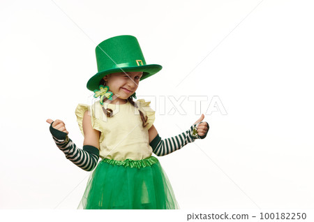 Adorable child, Irish little girl wearing clover leaf earrings, striped gloves, carnival green hat and skirt, winking and thumbing up, looking at camera on isolated white background. St. Patrick's Day 100182250