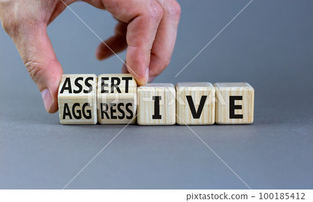 Aggressive or assertive symbol. Businessman turns wooden cubes, changes the word Aggressive to Assertive. Beautiful grey background, copy space. Business, psychological aggressive assertive concept. Aggressive or assertive symbol. Businessman turns wooden cubes, changes the word Aggressive to Assertive. Beautiful grey background, copy space. Business, psychological aggressive assertive concept. 100185412