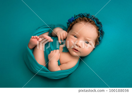 Top view of a newborn baby girl lying with her eyes open in a turquoise wrap with a turquoise headband and a flower on her head on a turquoise background. Portrait of a little girl 7 days, one week. 100188036