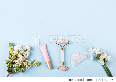 Pink jade roller and gua sha for face massage with cream or gel for the face and flowers daffodils and lilacs. Facial massager tools. Anti age, lifting and toning care. Modern selfcare concept. 100189486