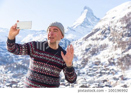 Man recording video with phone on background with Matterhorn mountain covered with snow on sunny day 100196992