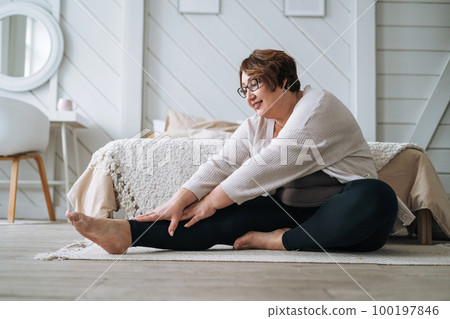 Middle aged plus size smiling brunette woman in home wear doing stretching in bedroom Middle aged plus size smiling brunette woman in home wear doing stretching in bedroom 100197846