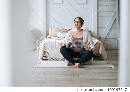 Middle aged plus size smiling brunette woman in home wear practicing yoga in bedroom Middle aged plus size smiling brunette woman in home wear practicing yoga in bedroom 100197847