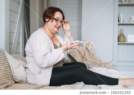 Smiling middle aged plus size woman using mobile phone on bed at home Smiling middle aged plus size woman using mobile phone on bed at home 100202292