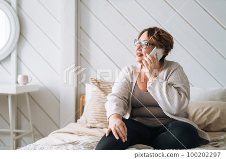 Smiling middle aged plus size woman using mobile phone on bed at home Smiling middle aged plus size woman using mobile phone on bed at home 100202297