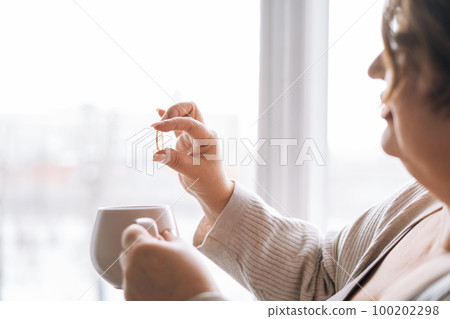 Middle aged woman holding omega 3 capsule and mug of water in hands near window at home Middle aged woman holding omega 3 capsule and mug of water in hands near window at home 100202298