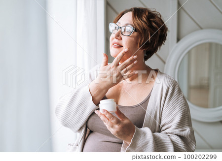 Smiling middle aged plus size woman with day cream near window at home Smiling middle aged plus size woman with day cream near window at home 100202299