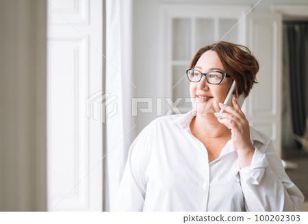Adult charming brunette woman in glasses plus size body positive using mobile near window Adult charming brunette woman in glasses plus size body positive using mobile near window 100202303