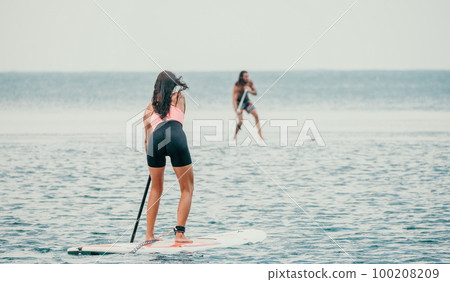 Sea woman and man on sup. Silhouette of happy young woman and man, surfing on SUP board, confident paddling through water surface. Idyllic sunset. Active lifestyle at sea or river. 100208209