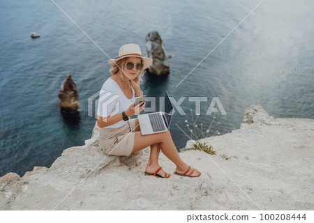 Freelance women sea working on the computer. Good looking middle aged woman typing on a laptop keyboard outdoors with a beautiful sea view. The concept of remote work. 100208444