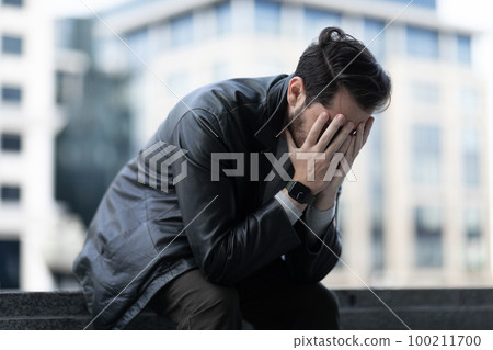 distressed middle-aged man crying with his eyes closed while sitting on the street 100211700