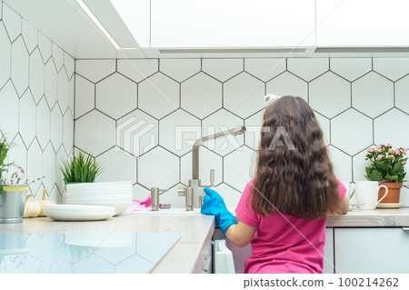 Little girl in protective gloves washing up dishes in kitchen sink. Portrait of child tidying up kitchen back view. 100214262