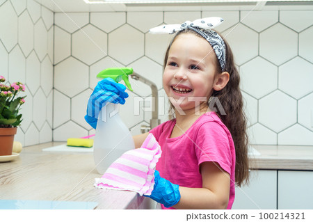 Joyful little girl in protective gloves wipe kitchen set with household rag and detergent sprayer. Home wet cleaning. Joyful little girl in protective gloves wipe kitchen set with household rag and detergent sprayer. Home wet cleaning. 100214321