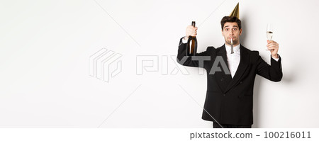 Concept of holidays and lifestyle. Handsome guy celebrating birthday, blowing party whistle and holding champagne, saying a toast, standing in suit over white background 100216011