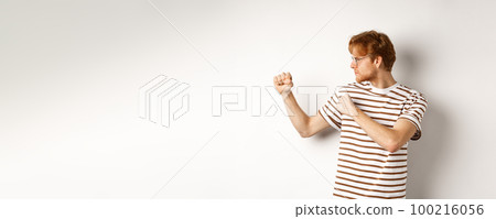Funny young man with red hair raising fists for fight, shadow boxing and looking serious at left side, standing over white background 100216056