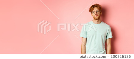 Image of young redhead man with beard, wearing t-shirt and glasses, looking left with bored unbothered face, standing over pink background 100216126