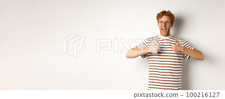 Young amazed man in red hair checking out something awesome, saying yes and showing thumbs-up, standing over white background 100216127
