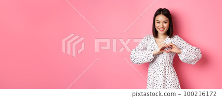 Romantic asian woman showing heart sign, I love you gesture, smiling cute at camera, standing in dress over pink background 100216172
