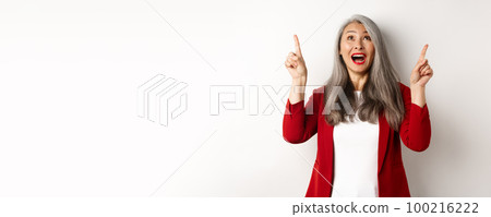 Happy asian lady in red blazer and makeup, looking and pointing fingers up, showing special offer, standing over white background Happy asian lady in red blazer and makeup, looking and pointing fingers up, showing special offer, standing over white background 100216222