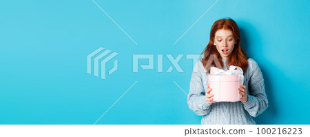 Beautiful redhead girl holding gift in pink cute box, looking at present with surprised face, standing over blue background 100216223