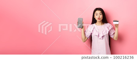Online shopping. Excited asian woman showing plastic credit card with empty smartphone screen, advertising internet shop, standing on pink background 100216236