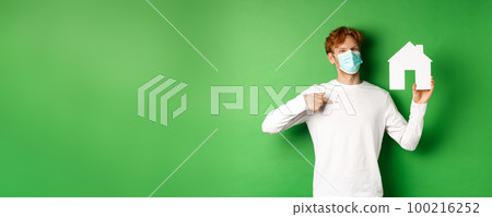 Real estate and covid-19 pandemic concept. Smiling redhead man in face mask pointing at paper house cutout, showing broker agency, standing over green background Real estate and covid-19 pandemic concept. Smiling redhead man in face mask pointing at paper house cutout, showing broker agency, standing over green background 100216252