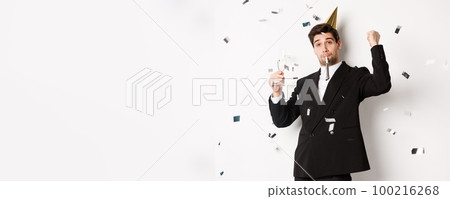 Handsome party guy in black suit having fun, celebrating new year, blowing whistle and drinking champagne while confetti falling, standing happy against white background Handsome party guy in black suit having fun, celebrating new year, blowing whistle and drinking champagne while confetti falling, standing happy against white background 100216268