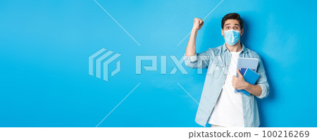 Education, covid-19 and social distancing. Excited male student in medical mask triumphing, raising fist up and holding notebooks, standing over blue background 100216269