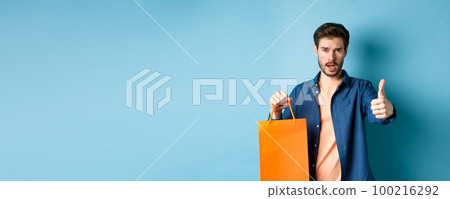 Cool guy showing thumb up and holding orange shopping bag, standing on blue background 100216292