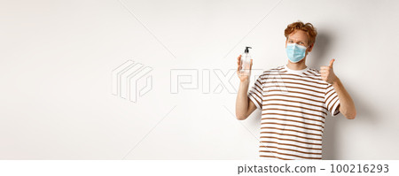 Covid-19, health and lifestyle concept. Smiling male model with red hair, wearing face mask, showing hand sanitizer and thumb-up, recommend to use antiseptic, white background 100216293