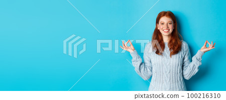 Smiling confident girl with red hair staying patient, holding hands in zen, meditation pose and staring at camera, practice yoga, standing calm against blue background Smiling confident girl with red hair staying patient, holding hands in zen, meditation pose and staring at camera, practice yoga, standing calm against blue background 100216310